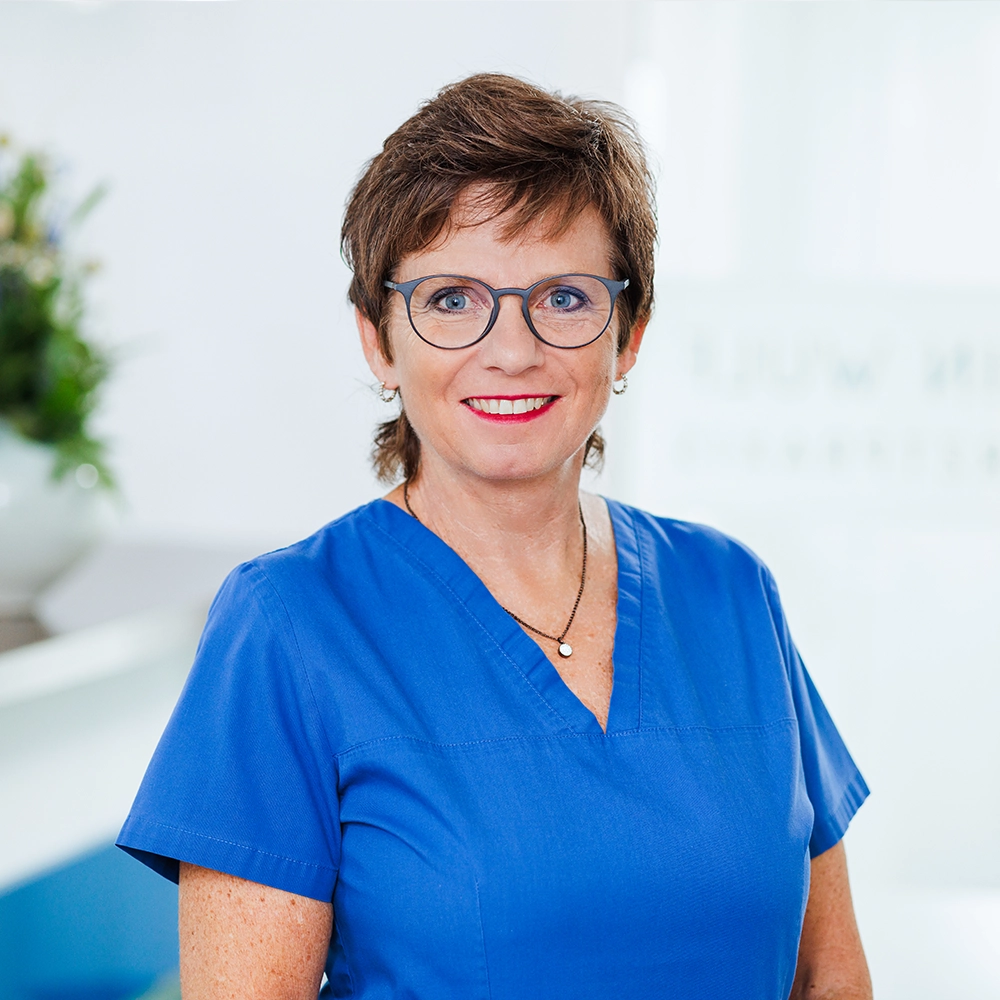 Mitarbeiterin Gisela mit Kurzhaarfrisur und Brille lächelt in blauer Praxiskleidung