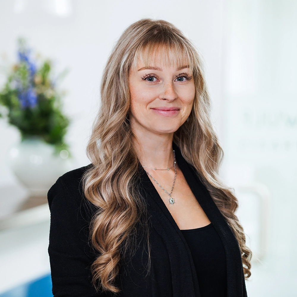 Mitarbeiterin Janet mit langen blonden Haaren lächelt freundlich vor hellem Praxishintergrund
